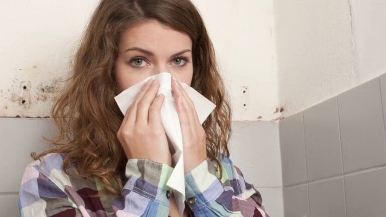 a woman covering her nose with tissue paper seems she is having flue because of the mold that can be seen behind her on the wall.